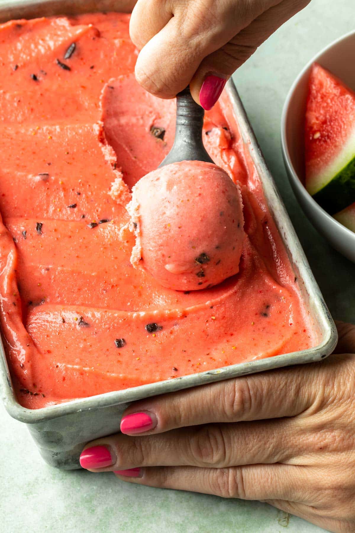 A hand scooping watermelon sorbet from a container, with a bowl of watermelon wedges in the background. 