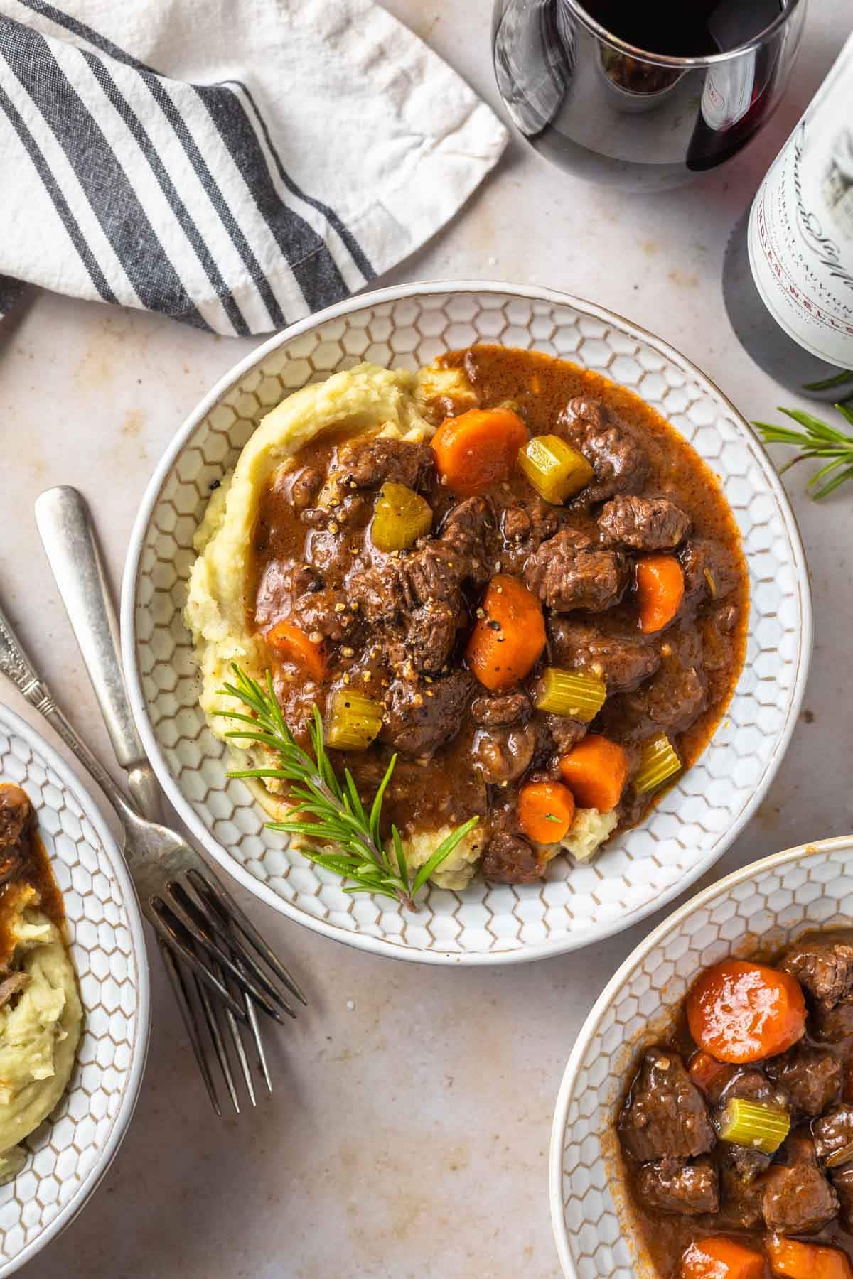 overhead view of red wine beef stew in a bowl over mashed potatoes garnished with rosemary on a counter top next to two other bowls and a glass of wine
