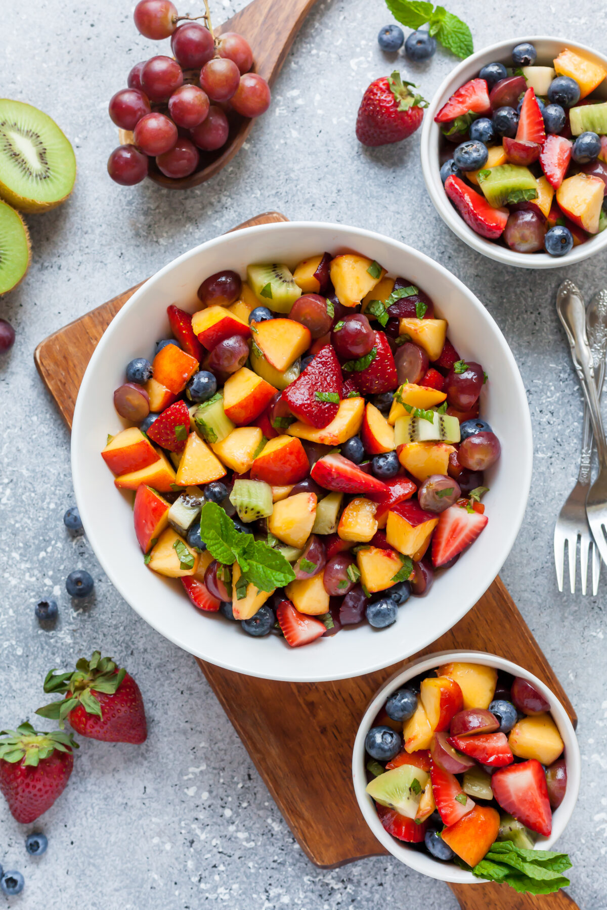A large bowl of vibrant summer fruit salad with strawberries, blueberries, grapes, peaches, and kiwi—garnished with mint leaves—sits on a wooden board, surrounded by smaller bowls of summer fruit salad and scattered fresh fruit.