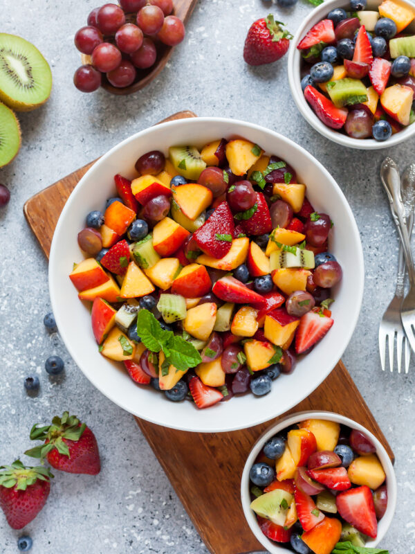 A large bowl of vibrant summer fruit salad with strawberries, blueberries, grapes, peaches, and kiwi—garnished with mint leaves—sits on a wooden board, surrounded by smaller bowls of summer fruit salad and scattered fresh fruit.
