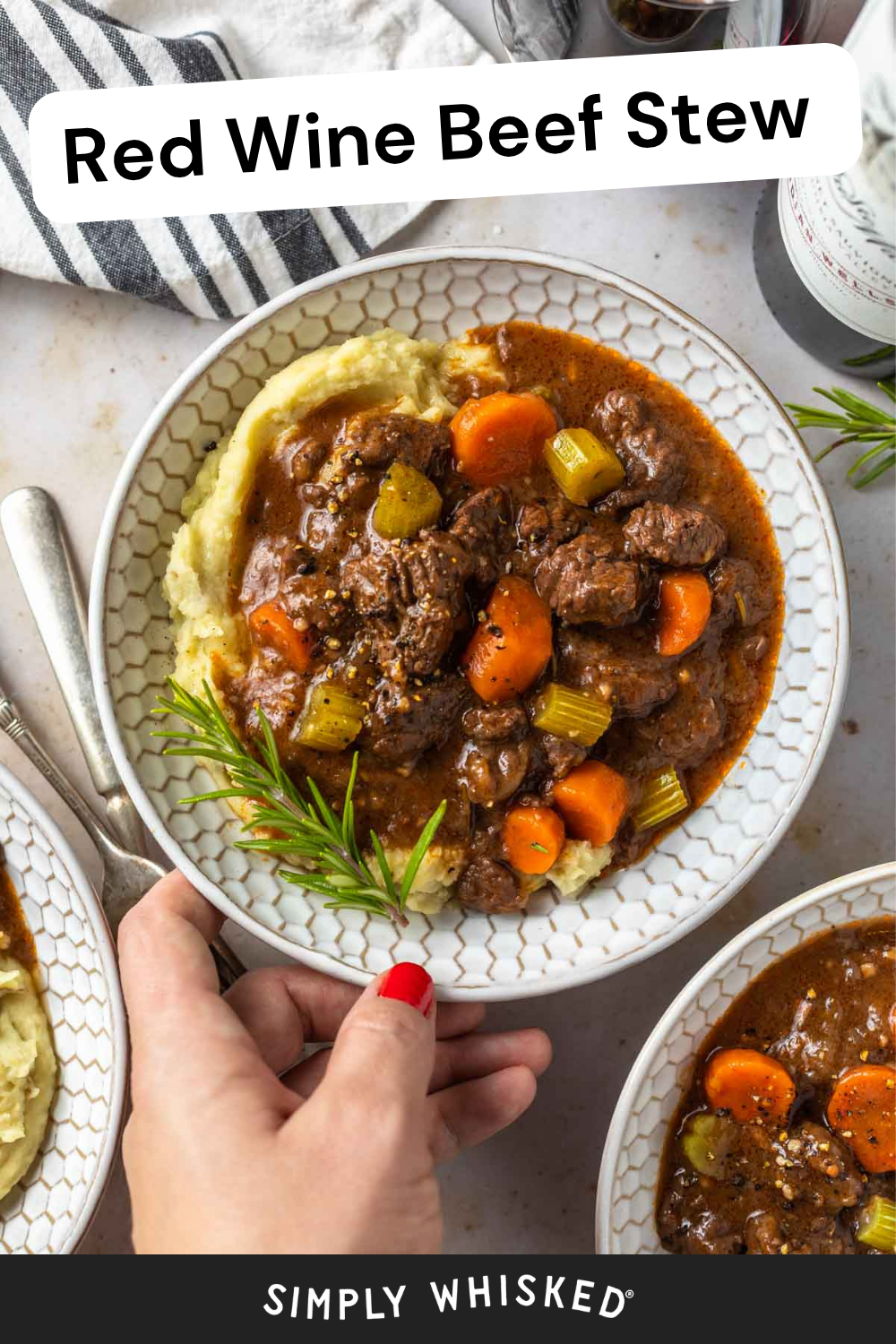 A hand holding a bowl of hearty red wine beef stew with carrots and celery, served over creamy mashed potatoes and garnished with fresh rosemary. Other bowls of red wine beef stew and a striped napkin are nearby.