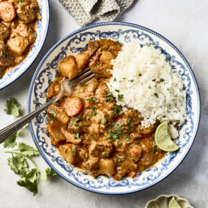 A decorative plate with white rice and slow cooker massaman curry featuring chunks of meat, vegetables, and chopped cilantro, served with lime wedges. A fork rests on the plate, and a cloth napkin is nearby.