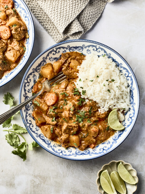 A decorative plate with white rice and slow cooker massaman curry featuring chunks of meat, vegetables, and chopped cilantro, served with lime wedges. A fork rests on the plate, and a cloth napkin is nearby.