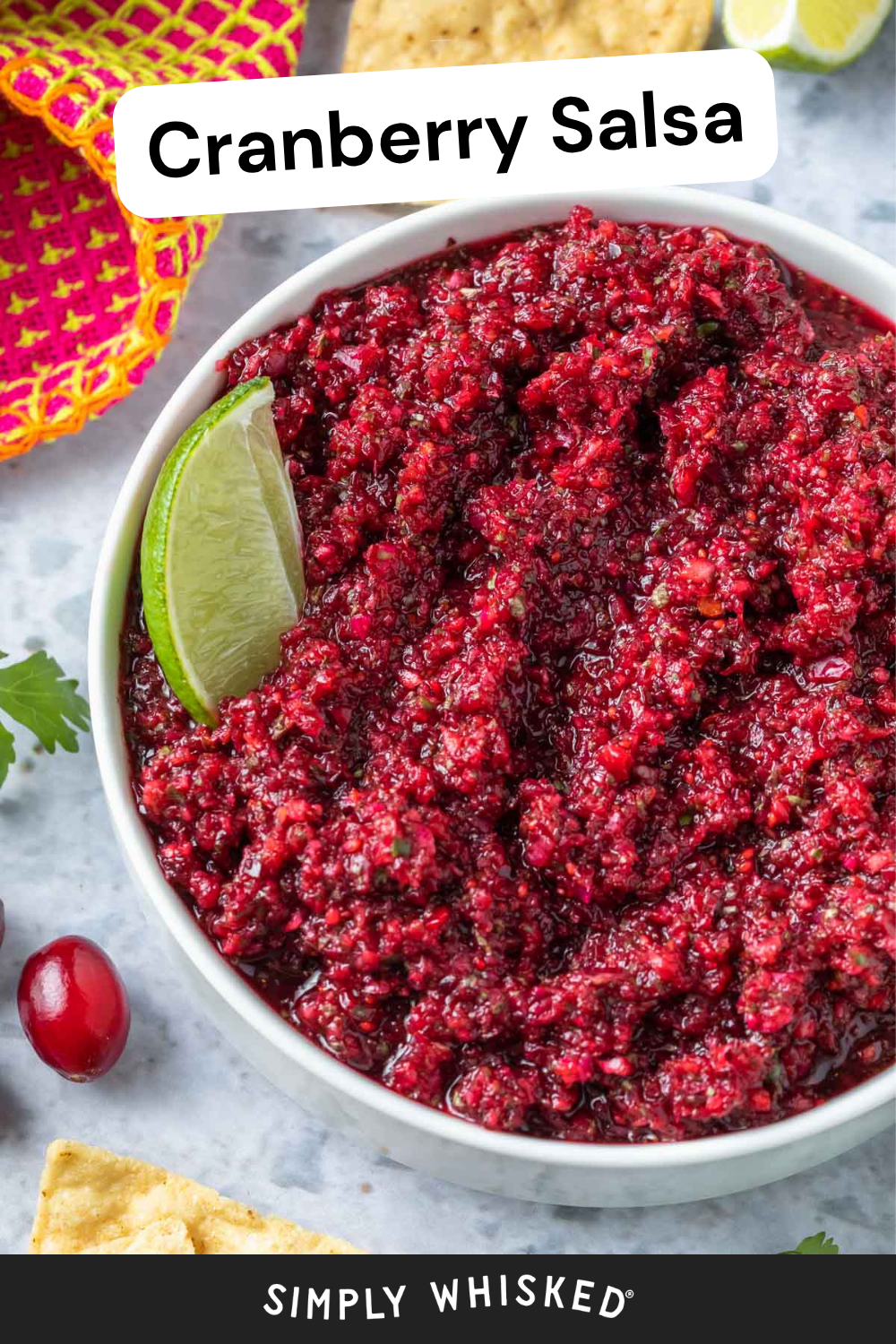 A bowl of cranberry salsa garnished with a lime wedge sits surrounded by chips, a fresh cranberry, and a colorful napkin. The words Cranberry Salsa are displayed at the top of the image, highlighting this vibrant and festive appetizer.