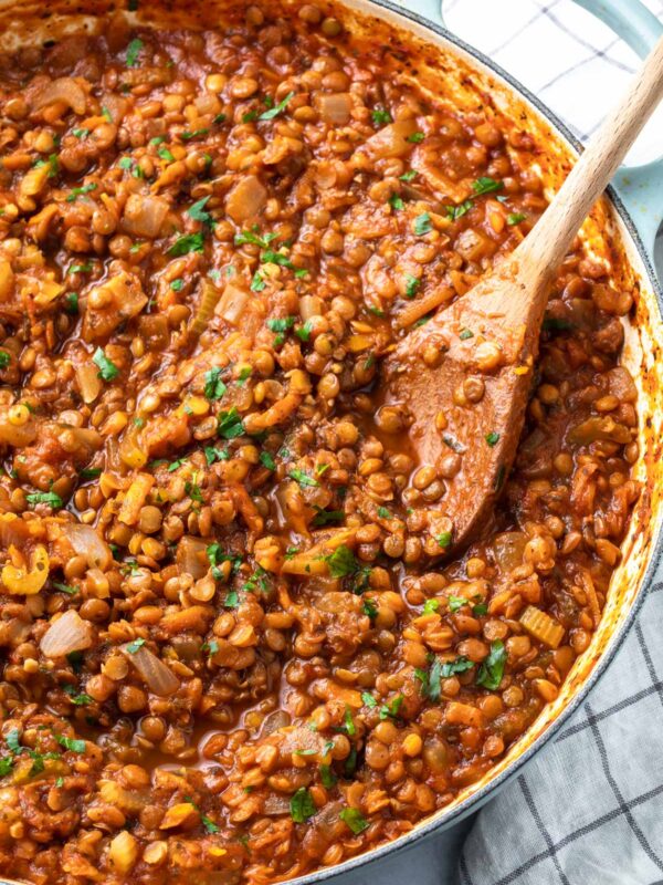 A close-up of a pot filled with hearty lentil bolognese stew in a tomato-based sauce, garnished with chopped parsley and stirred with a wooden spoon. The pot rests on a checkered kitchen towel.