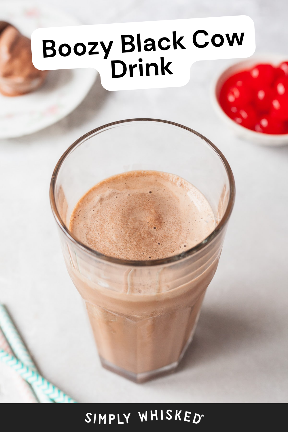 A glass of chocolate milkshake labeled Black Cow Drink sits on a light surface near a bowl of cherries, a plate with ice cream, and two striped straws.