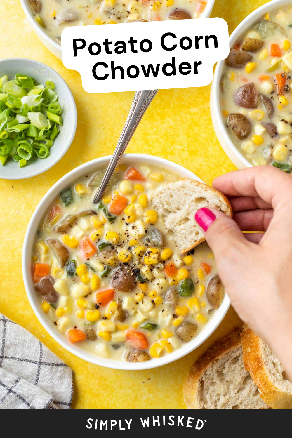 A hand dips bread into a bowl of hearty potato corn chowder, brimming with visible corn, carrots, potatoes, and fresh herbs. Sliced green onions and extra bread are nearby on a cheerful yellow surface.