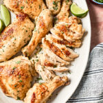A platter with coconut chicken pieces, including sliced breast, thighs, and drumsticks, garnished with chopped herbs and served with lime wedges. A bowl of herbs and a striped napkin are nearby.