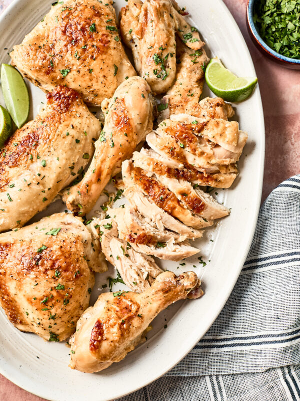 A platter with coconut chicken pieces, including sliced breast, thighs, and drumsticks, garnished with chopped herbs and served with lime wedges. A bowl of herbs and a striped napkin are nearby.