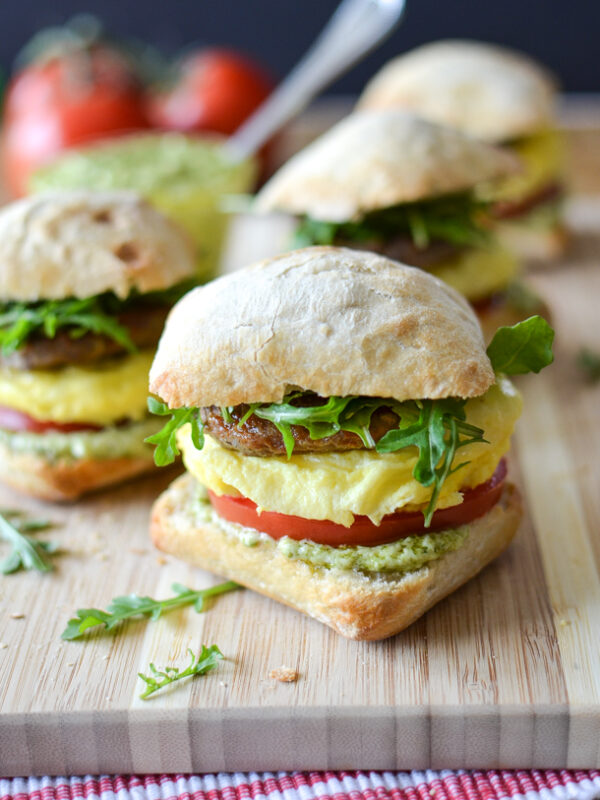 Breakfast sandwiches featuring sausage and scrambled egg sandwich options, plus tomato slices, arugula, and veggie patties on ciabatta rolls are arranged on a wooden board with a red and white towel and fresh tomatoes in the background.