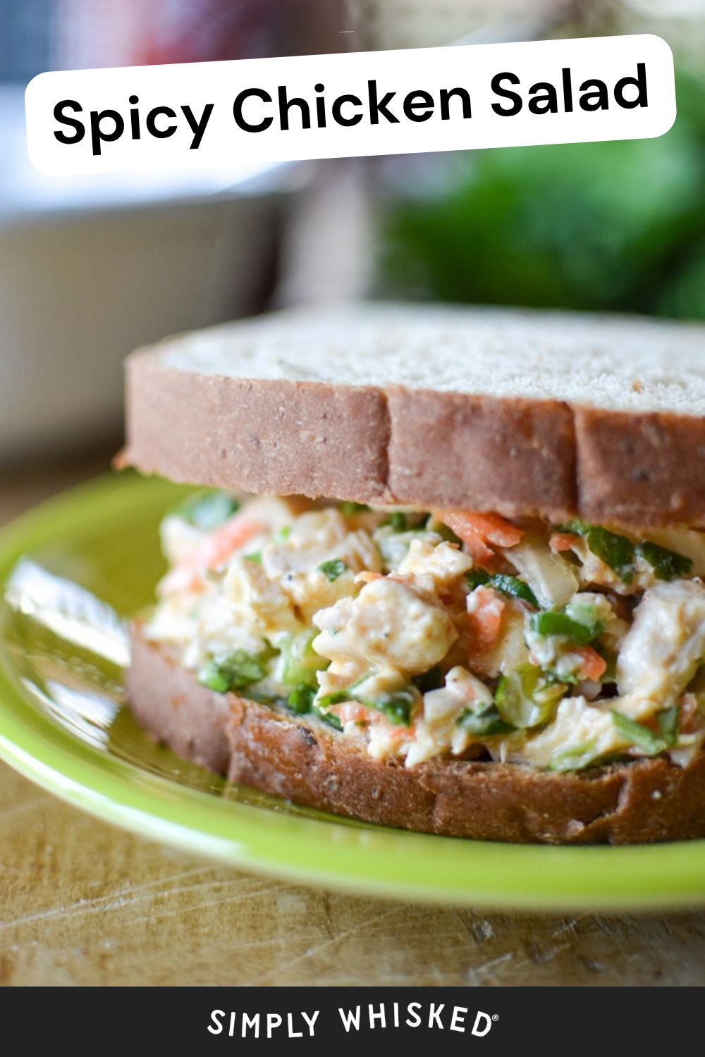 A spicy chicken salad sandwich on whole-wheat bread sits on a bright green plate. The filling contains chunks of spicy chicken salad, diced carrots, and green herbs. Spicy Chicken Salad text is at the top; Simply Whisked logo is at the bottom.