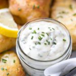 close up of homemade tartar sauce in a small mason jar plated with fried cod and lemon slices in a parchment lined cast iron skillet