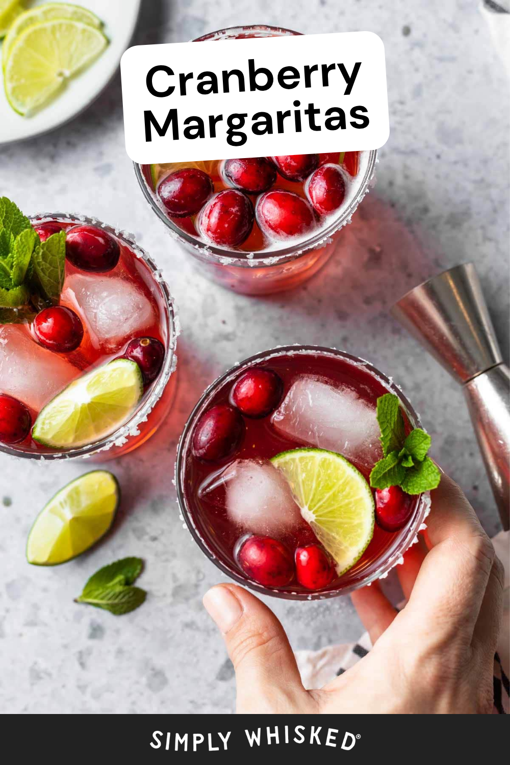 Three cranberry margaritas with ice, cranberries, mint, and lime wedges on a gray surface. A hand holds one glass, ready to toast. Lime slices and a metal jigger are nearby, adding to the festive setup. Text reads Cranberry Margaritas and Simply Whisked—perfect inspiration for your next batch of homemade cranberry margaritas.