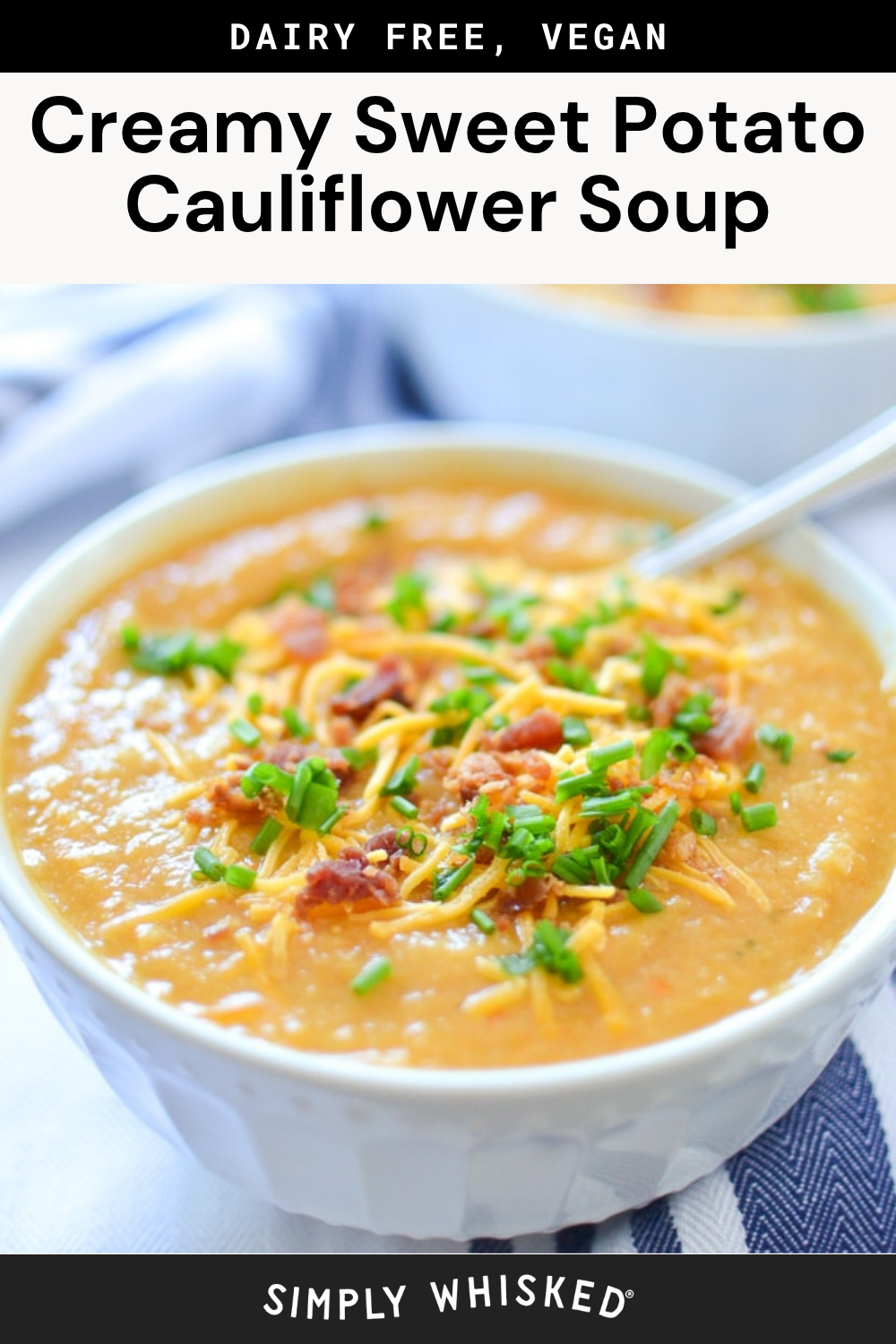 A bowl of creamy orange cauliflower potato soup topped with chopped chives, shredded cheese, and bits of what appears to be bacon, with a spoon in the bowl. A second bowl is blurred in the background.