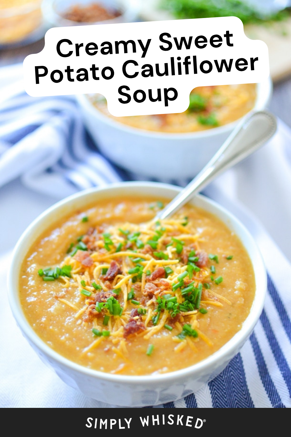 A bowl of creamy cauliflower potato soup topped with shredded cheese, bacon bits, and chopped chives, with a spoon and a striped napkin on the side.