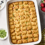 A breakfast casserole topped with golden brown tater tots, garnished with chopped green onions, sits in a white baking dish. Strawberries, extra green onions, plates, and a yellow-checkered cloth surround it.
