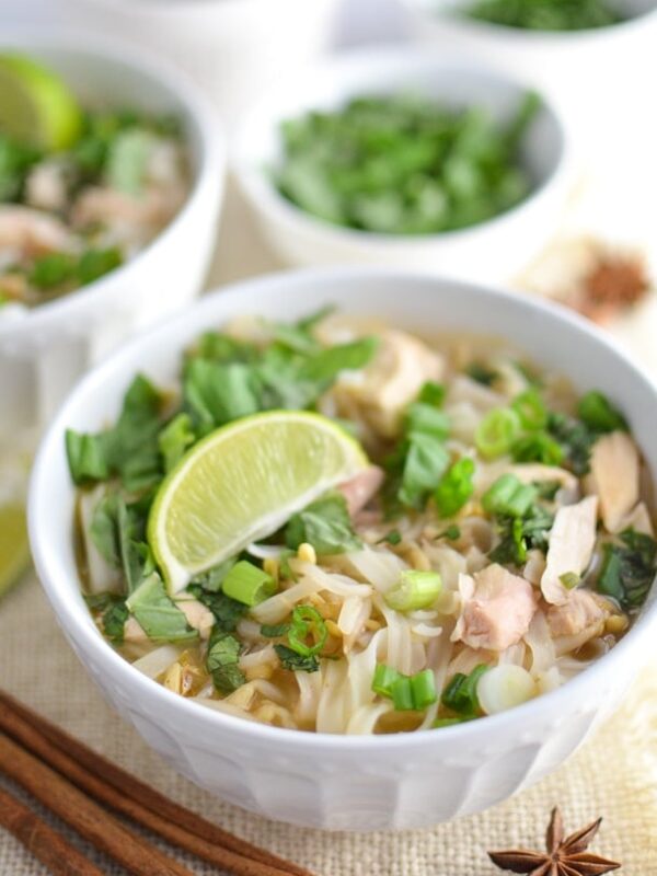 A white bowl of chicken pho topped with fresh herbs, chopped green onions, and a lime wedge. Cinnamon sticks and a star anise sit in the foreground, with bowls of herbs in the background completing this inviting chicken pho scene.