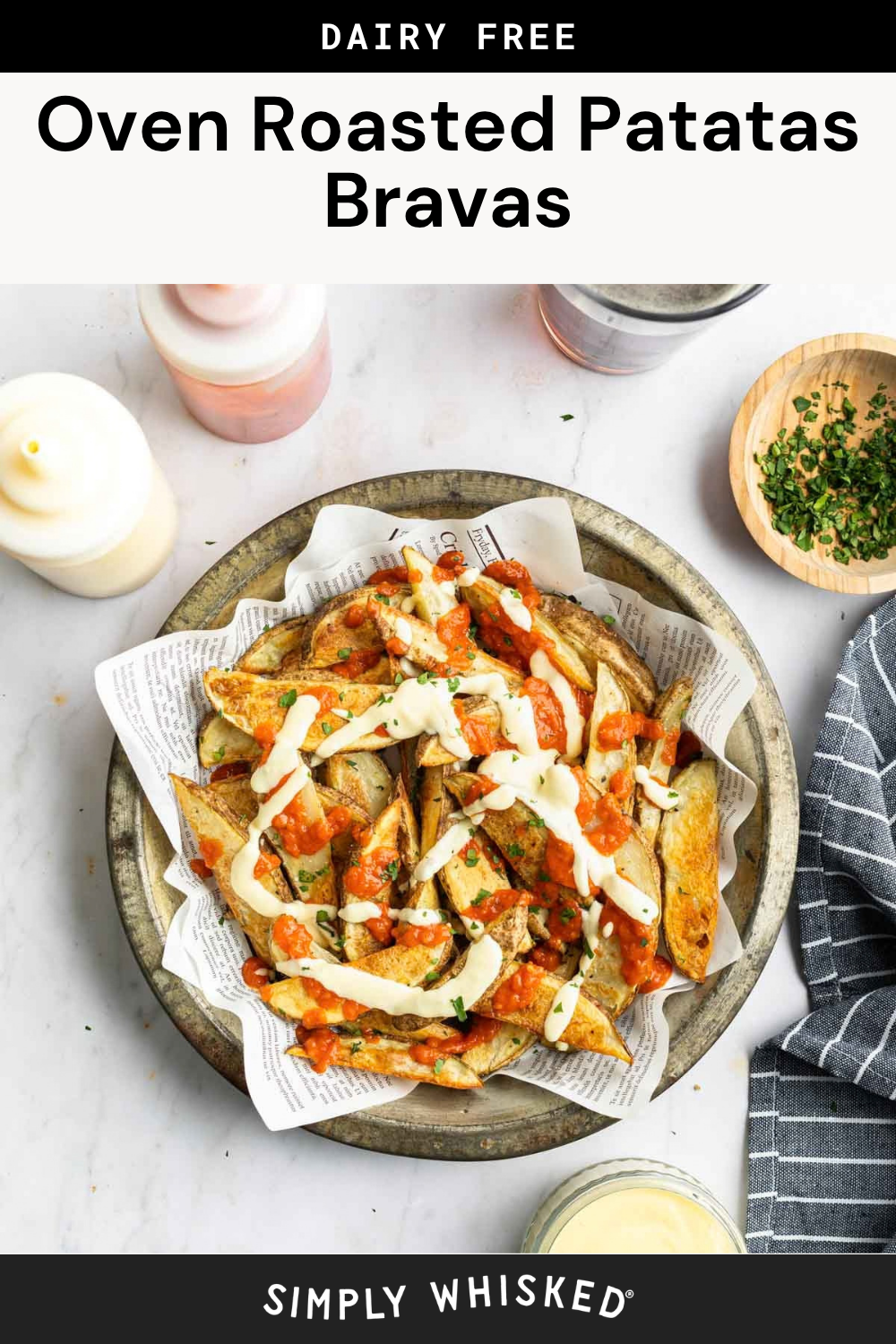 A plate of oven-roasted patatas bravas topped with vibrant red and white sauces, served on newspaper in a metal dish, surrounded by sauce bottles, fresh herbs, and a striped napkin.