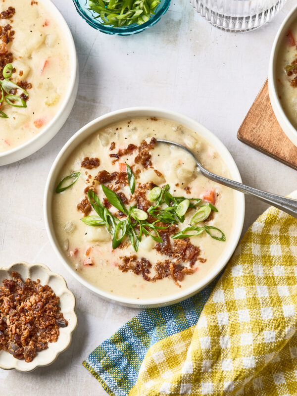 A bowl of creamy dairy free potato soup topped with chopped green onions and crispy bits, with a spoon inside. Surrounding the bowl are extra crispy bits, a small bowl of green onions, a glass of water, and a yellow checkered napkin.