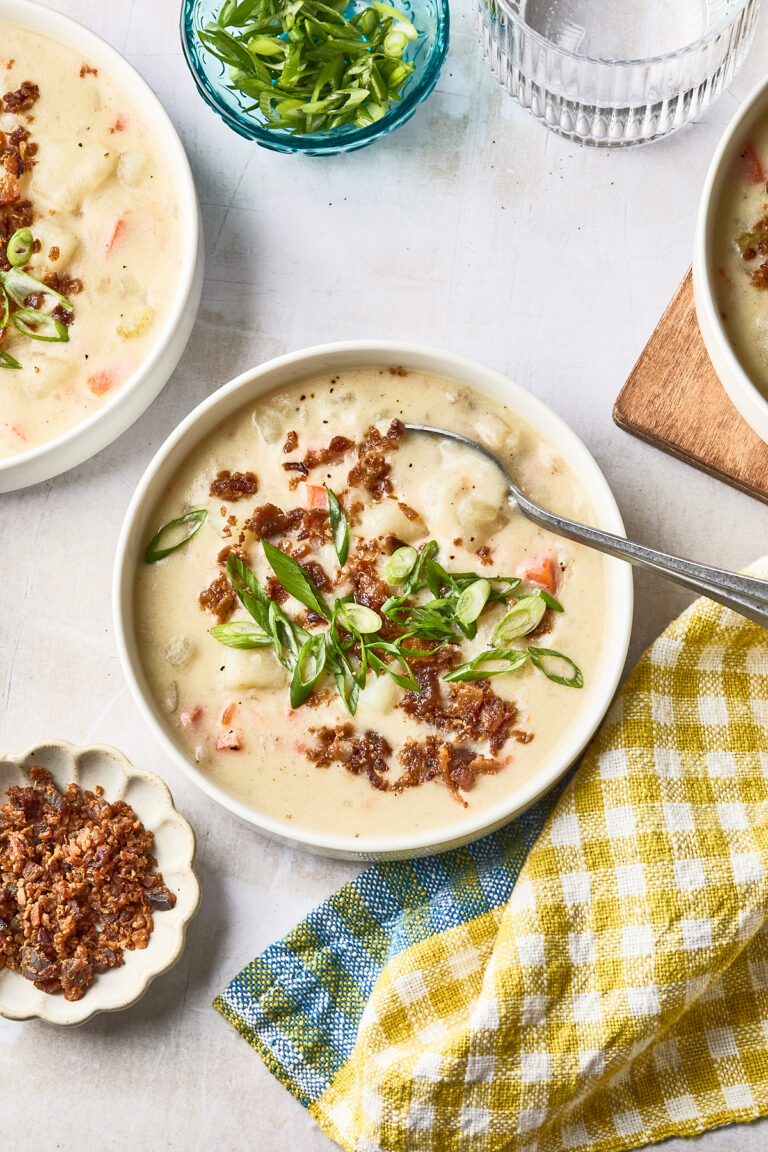 A bowl of creamy dairy free potato soup topped with chopped green onions and crispy bits, with a spoon inside. Surrounding the bowl are extra crispy bits, a small bowl of green onions, a glass of water, and a yellow checkered napkin.