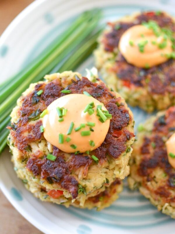 Three golden-brown tuna cakes topped with a dollop of orange sauce and chopped chives, served on a white plate with green stripes and garnished with a bundle of fresh chives.