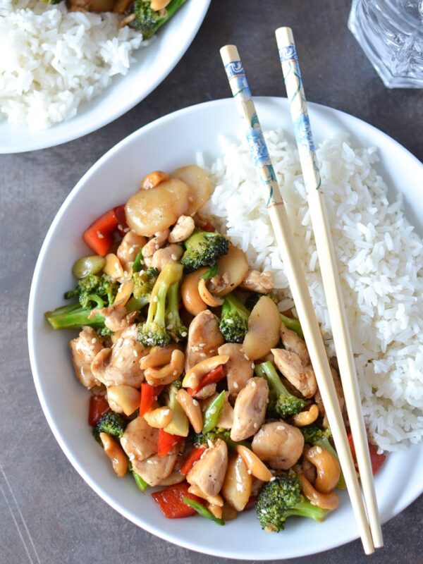 A white plate with white rice and Chinese cashew chicken—stir-fried chicken, broccoli, red peppers, water chestnuts, and cashews. Two decorated chopsticks rest on the side of the plate. Another plate and a glass of water are nearby.
