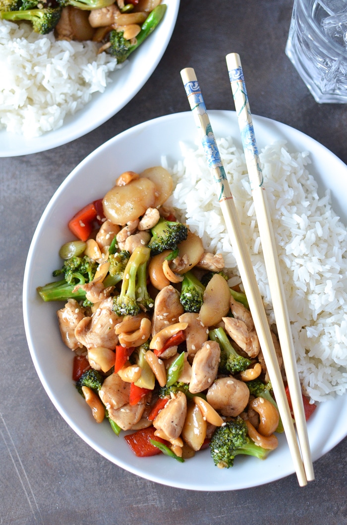 Cashew chicken in a bowl with white rice, with chopsticks resting across the bowl. 