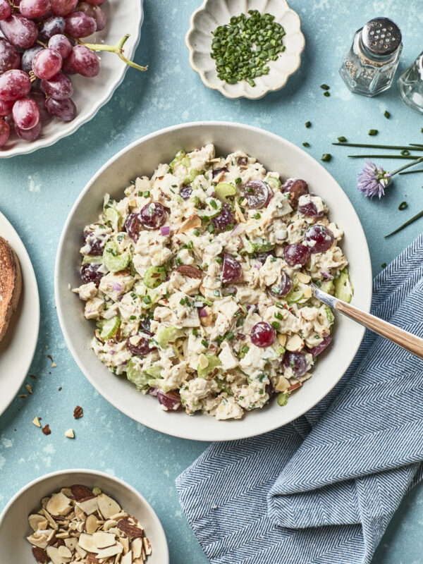 A bowl of chicken salad with grapes, sliced celery, and fresh herbs sits on a blue surface, surrounded by a plate of grapes, chopped chives, sliced almonds, and a salt and pepper shaker.
