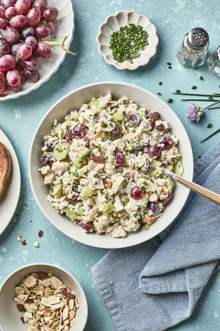 A bowl of chicken salad with grapes, sliced celery, and fresh herbs sits on a blue surface, surrounded by a plate of grapes, chopped chives, sliced almonds, and a salt and pepper shaker.