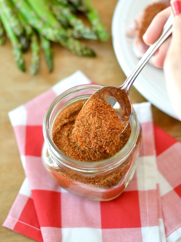 A hand holds a spoon scooping an easy BBQ dry rub from a glass jar, which sits on a red and white checkered cloth. Fresh asparagus and a white plate are in the background.