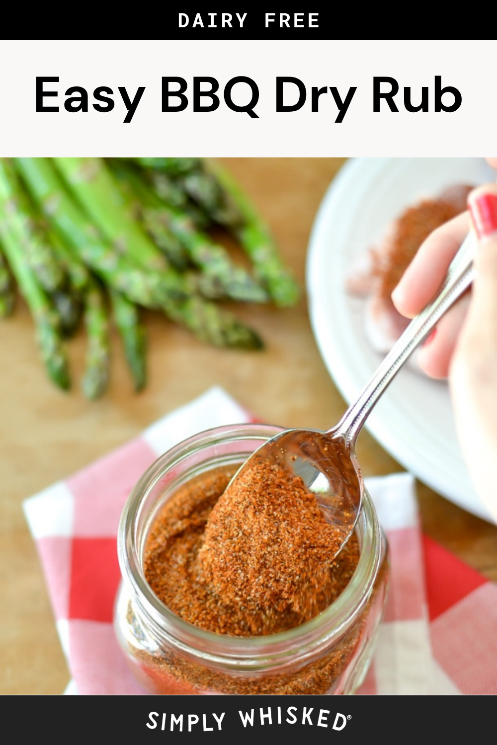 A hand scoops easy BBQ dry rub from a glass jar with a spoon. Fresh asparagus and a plate with more seasoning are in the background. Text reads “Dairy Free Easy BBQ Dry Rub.”.