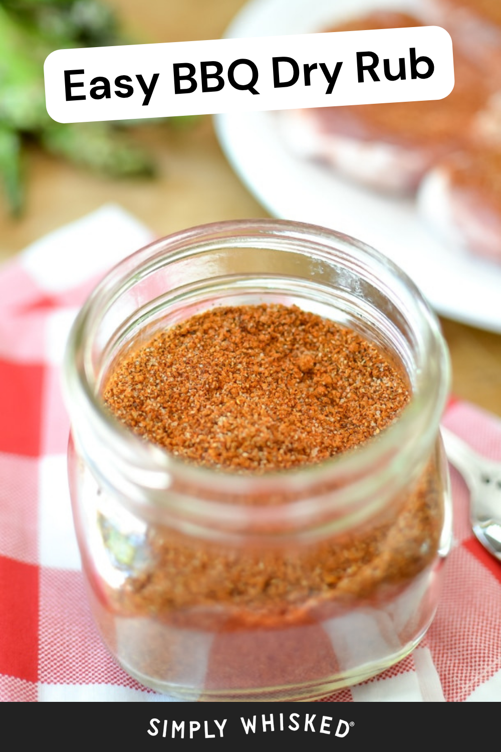 A glass jar filled with easy BBQ dry rub sits on a red and white cloth, with raw meat and herbs blurred in the background. The text reads “Easy BBQ Dry Rub.”.
