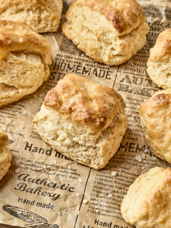 Golden brown dairy free biscuits are arranged on rustic parchment paper with bakery-themed text and designs. The biscuits look fluffy and freshly baked.