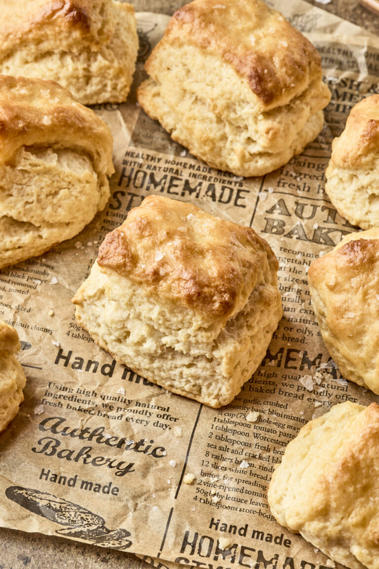 Golden brown dairy free biscuits are arranged on rustic parchment paper with bakery-themed text and designs. The biscuits look fluffy and freshly baked.