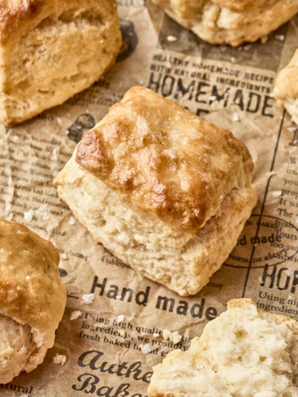 A close-up of golden, flaky homemade dairy free biscuits resting on brown parchment paper printed with words like hand made and authentic bakery. One biscuit is split open, revealing its soft, fluffy interior.