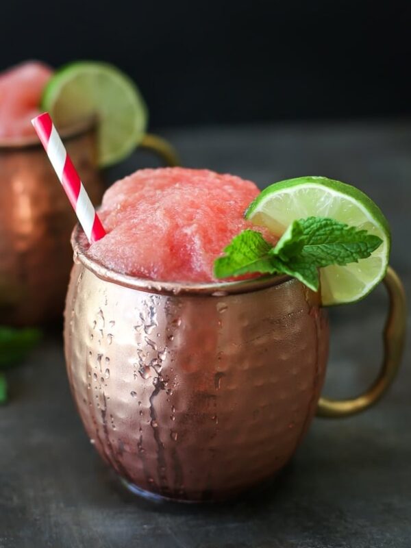 A copper mug filled with a frozen watermelon Moscow mule, garnished with a lime slice and fresh mint, with a red-and-white striped straw. Another similar drink is blurred in the background.