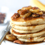 A stack of bananas foster pancakes topped with caramelized banana slices, pecans, and syrup sits on a white plate with a fork; a banana and a bowl of pecans are blurred in the background.