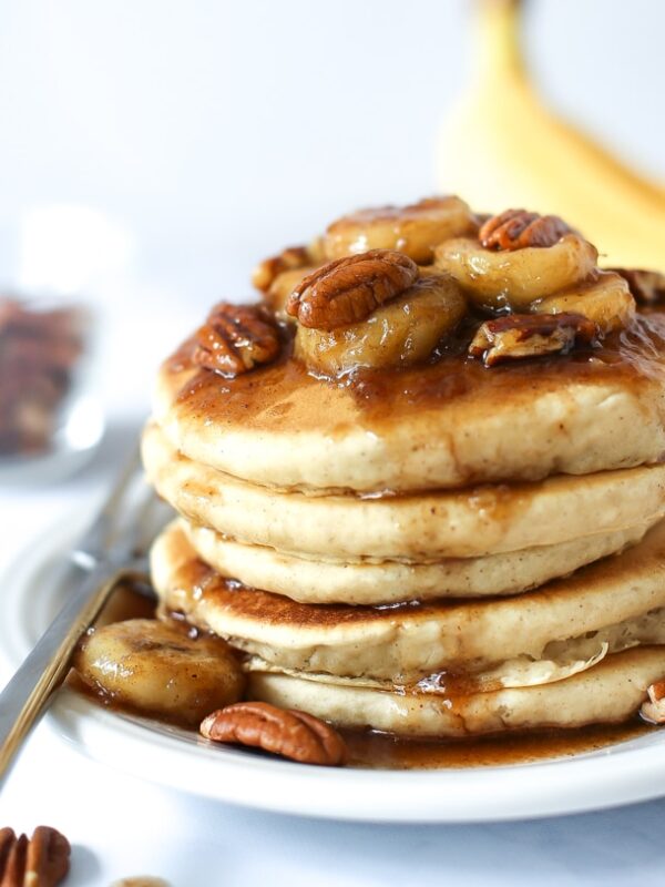 A stack of bananas foster pancakes topped with caramelized banana slices, pecans, and syrup sits on a white plate with a fork; a banana and a bowl of pecans are blurred in the background.