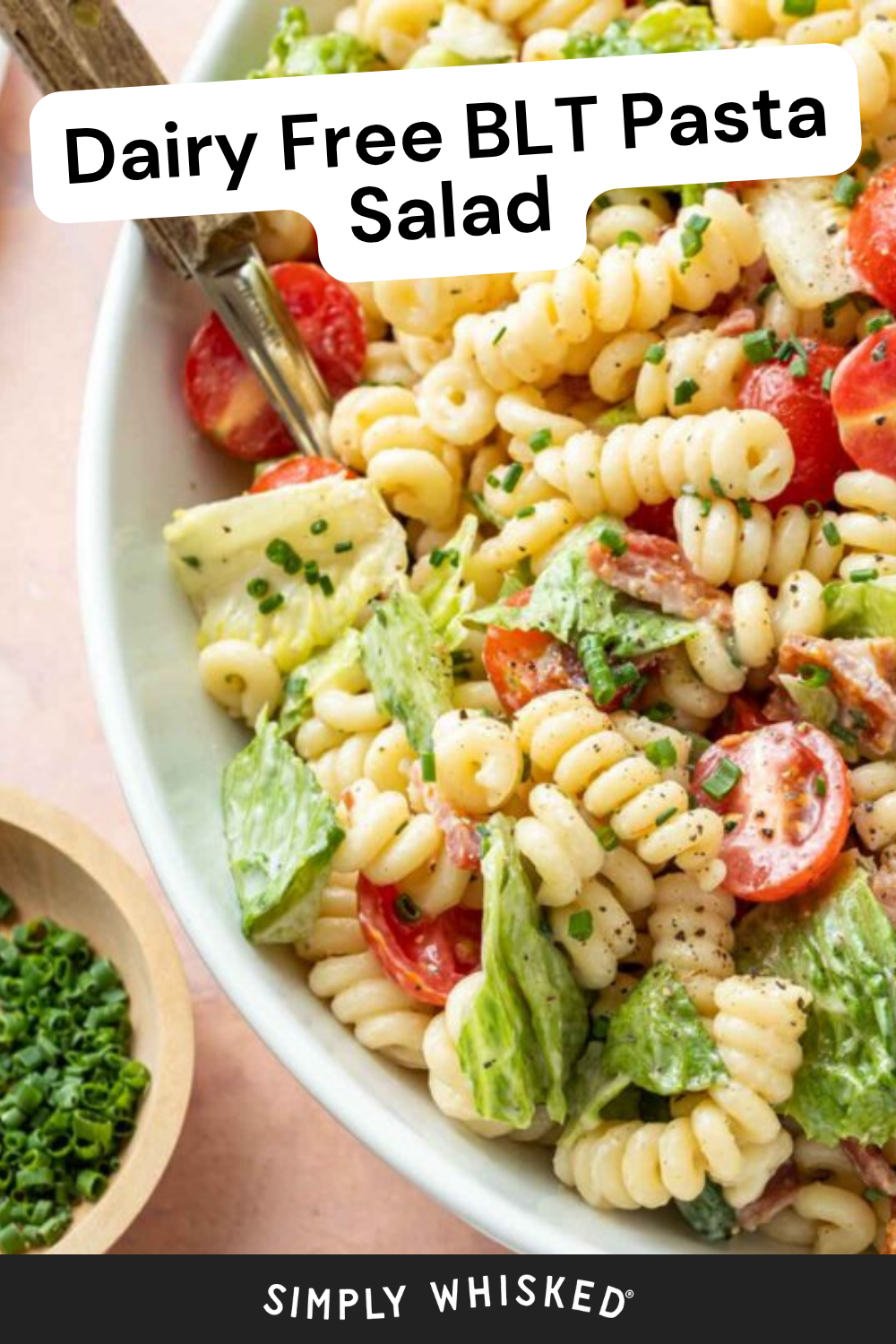 A bowl of BLT pasta salad with rotini pasta, lettuce, cherry tomatoes, avocado, and chopped chives sits ready to enjoy, accompanied by a fork and a small bowl of chives on the side. Text reads Dairy Free BLT Pasta Salad.