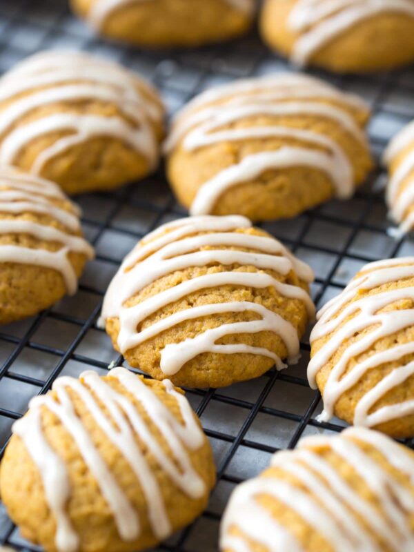 Soft pumpkin cookies with a white icing drizzle are arranged on a cooling rack. The golden-brown, round treats are evenly spaced across the wire grid, highlighting their tender texture.