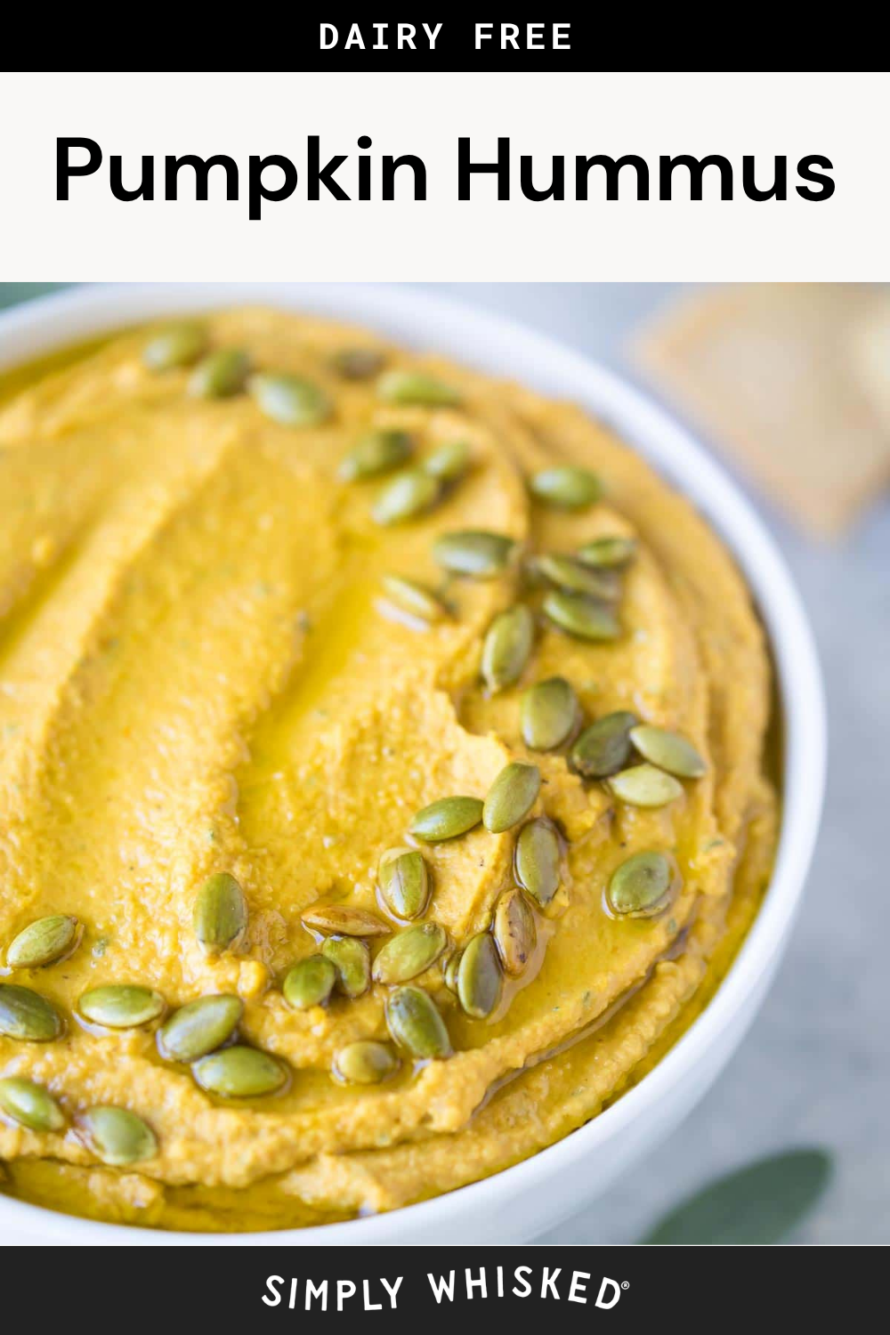 A bowl of creamy pumpkin hummus topped with green pumpkin seeds, with a piece of pita bread in the background. The text reads Dairy Free Roasted Garlic Pumpkin Hummus and Simply Whisked.