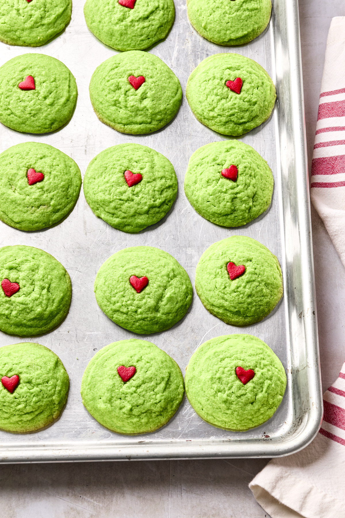 A baking tray filled with dairy free Grinch cookies, each topped with a small red heart decoration. A red-striped towel is partially visible on the right side of the image.