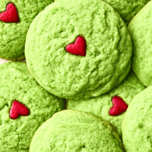 A close-up of dairy free Grinch cookies, topped with small red heart decorations, arranged in a pile on a white patterned surface.