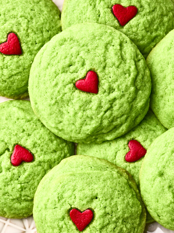 A close-up of dairy free Grinch cookies, topped with small red heart decorations, arranged in a pile on a white patterned surface.