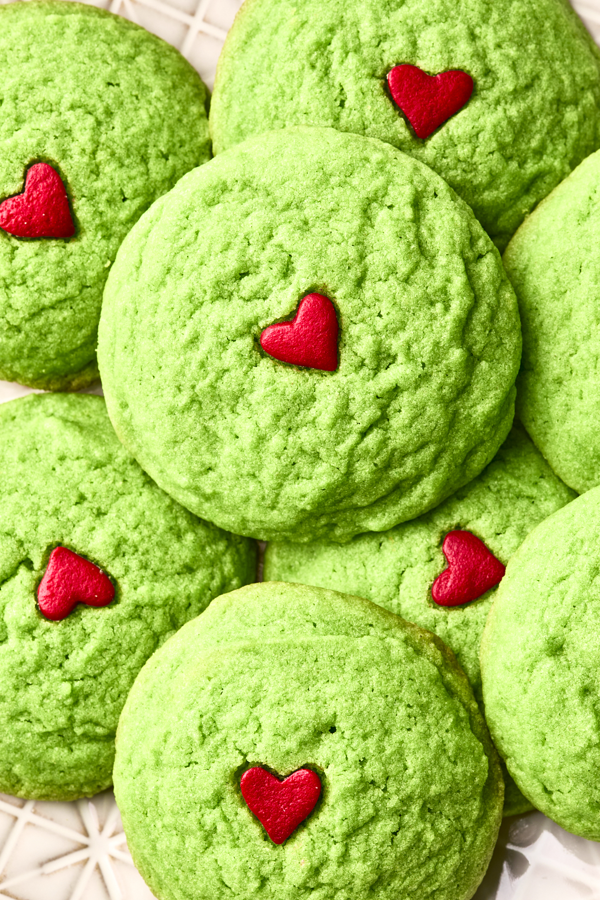 A close-up of dairy free Grinch cookies, topped with small red heart decorations, arranged in a pile on a white patterned surface.