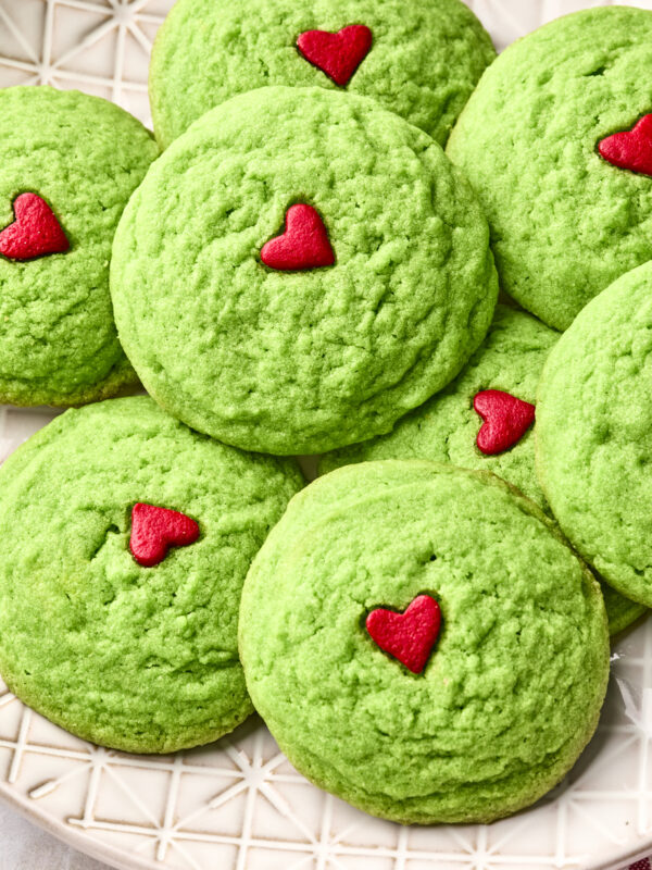 A plate of dairy free Grinch cookies, each vibrant green and topped with a small red heart in the center, arranged closely together on a patterned white plate.
