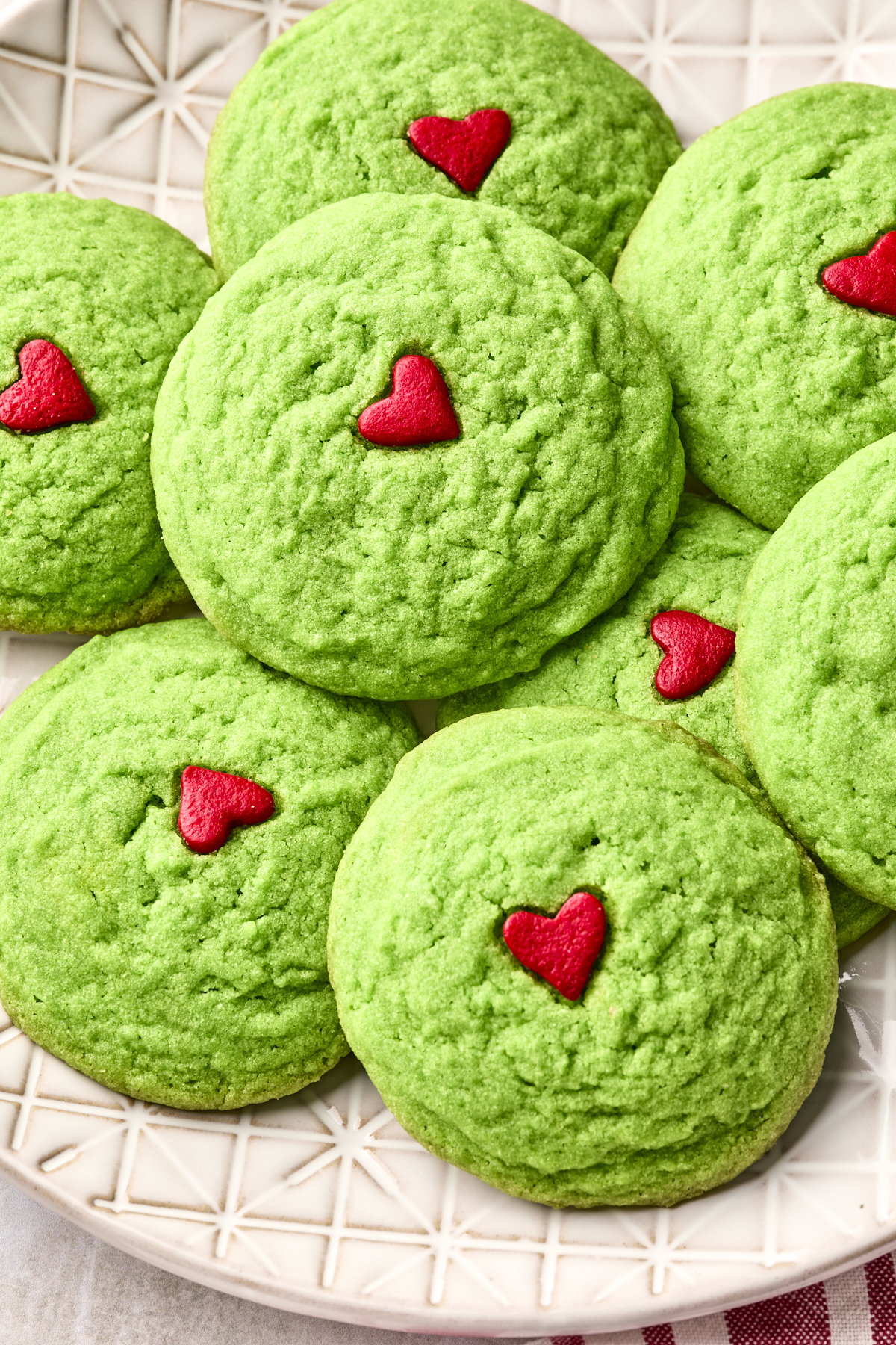 A plate of dairy free Grinch cookies, each vibrant green and topped with a small red heart in the center, arranged closely together on a patterned white plate.