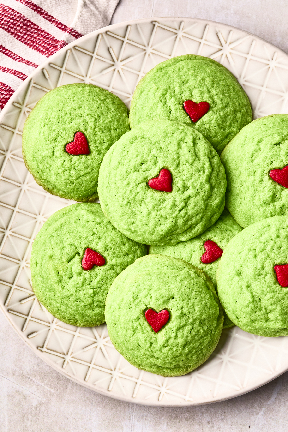A plate of round green Grinch cookies, each topped with a small red heart decoration, arranged on a patterned cream-colored plate. A red-and-white striped cloth is partially visible in the corner. Perfect dairy free Grinch cookies for any festive occasion!.