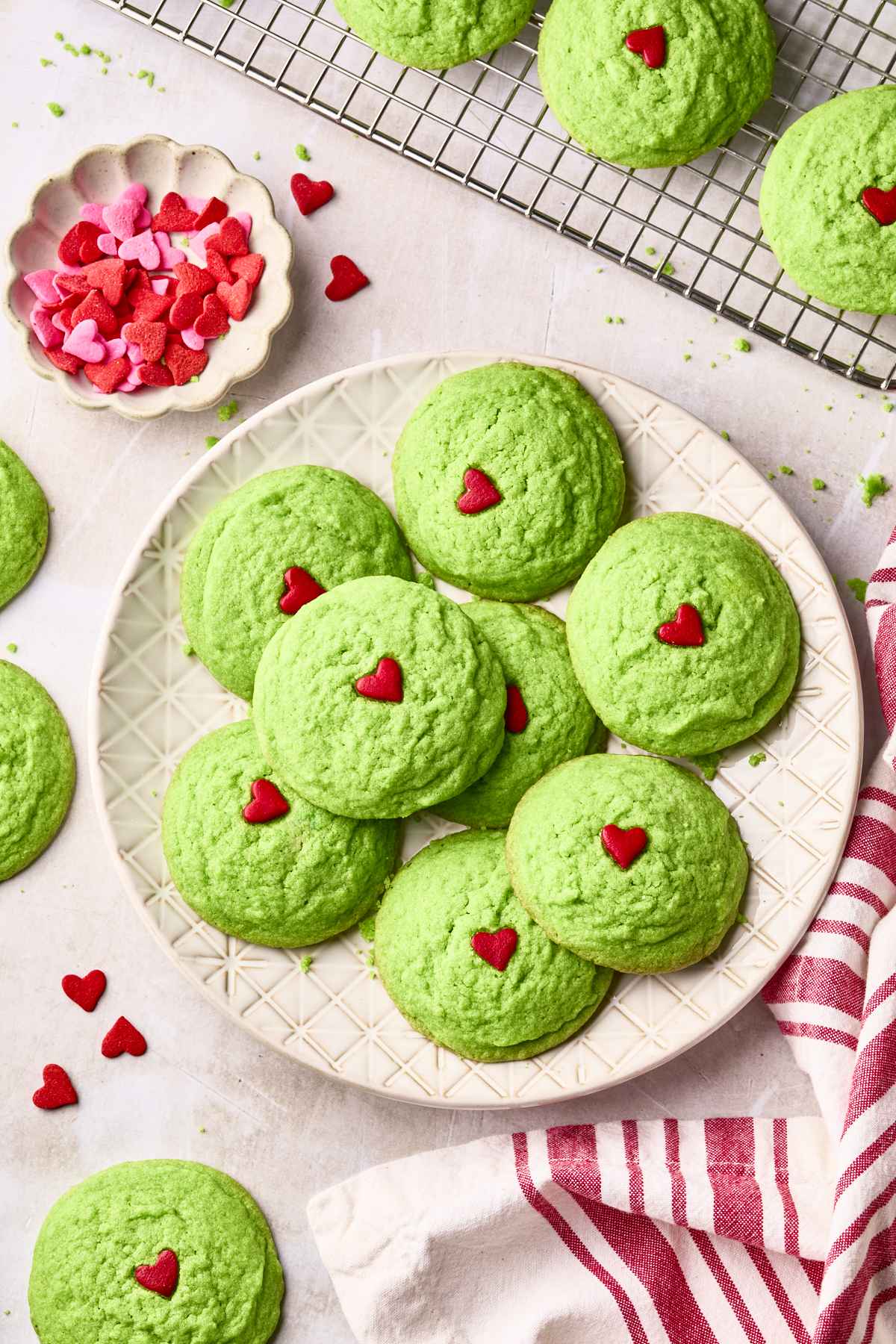 A plate of green Grinch cookies topped with red heart-shaped sprinkles, surrounded by more cookies, a bowl of heart sprinkles, and a red-and-white striped towel on a light surface.