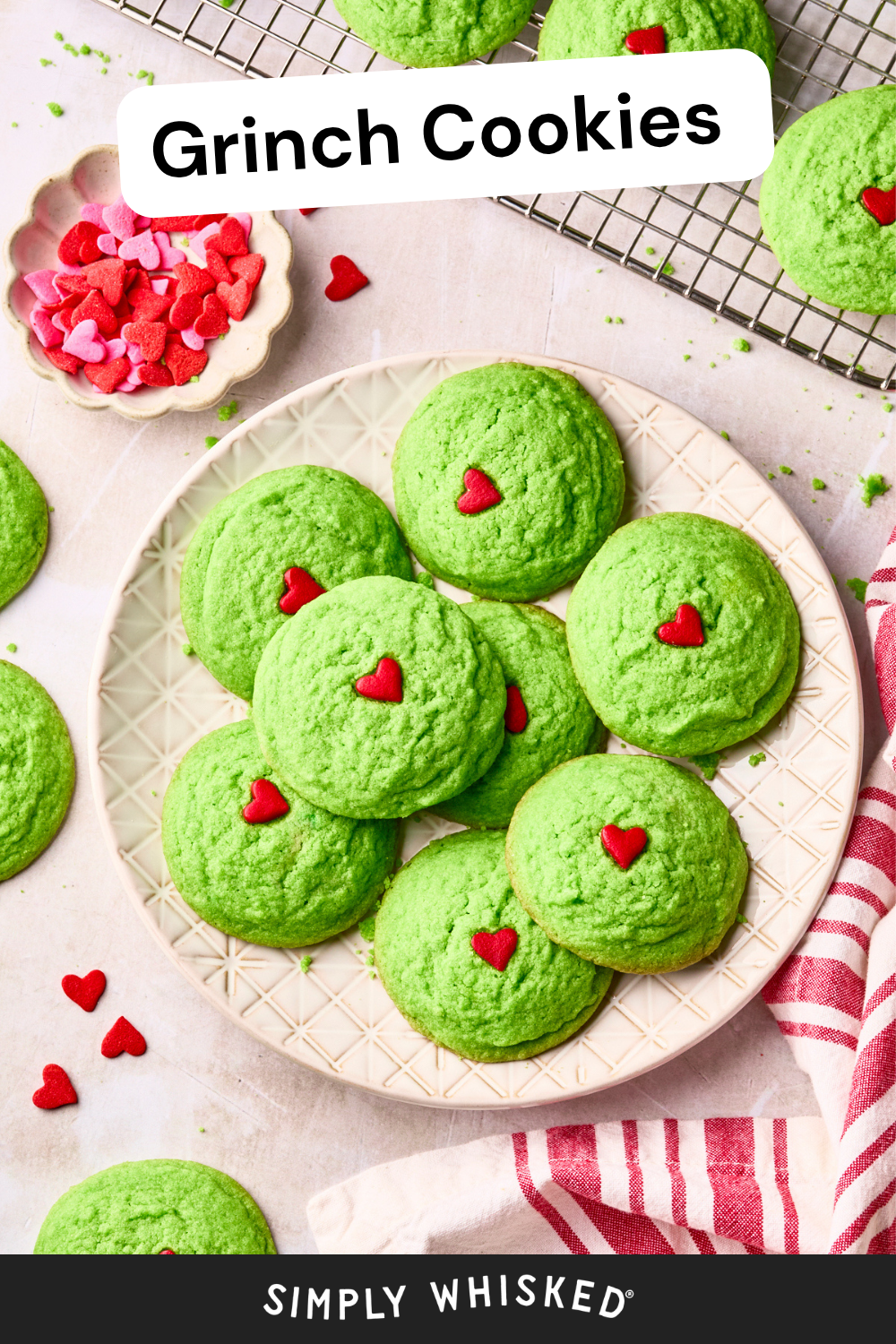 A plate of bright green Grinch cookies decorated with red heart sprinkles sits on a white surface. More cookies and red heart sprinkles are scattered nearby, with “Grinch Cookies” text at the top—perfect for anyone craving fun and festive dairy free Grinch cookies this holiday season.